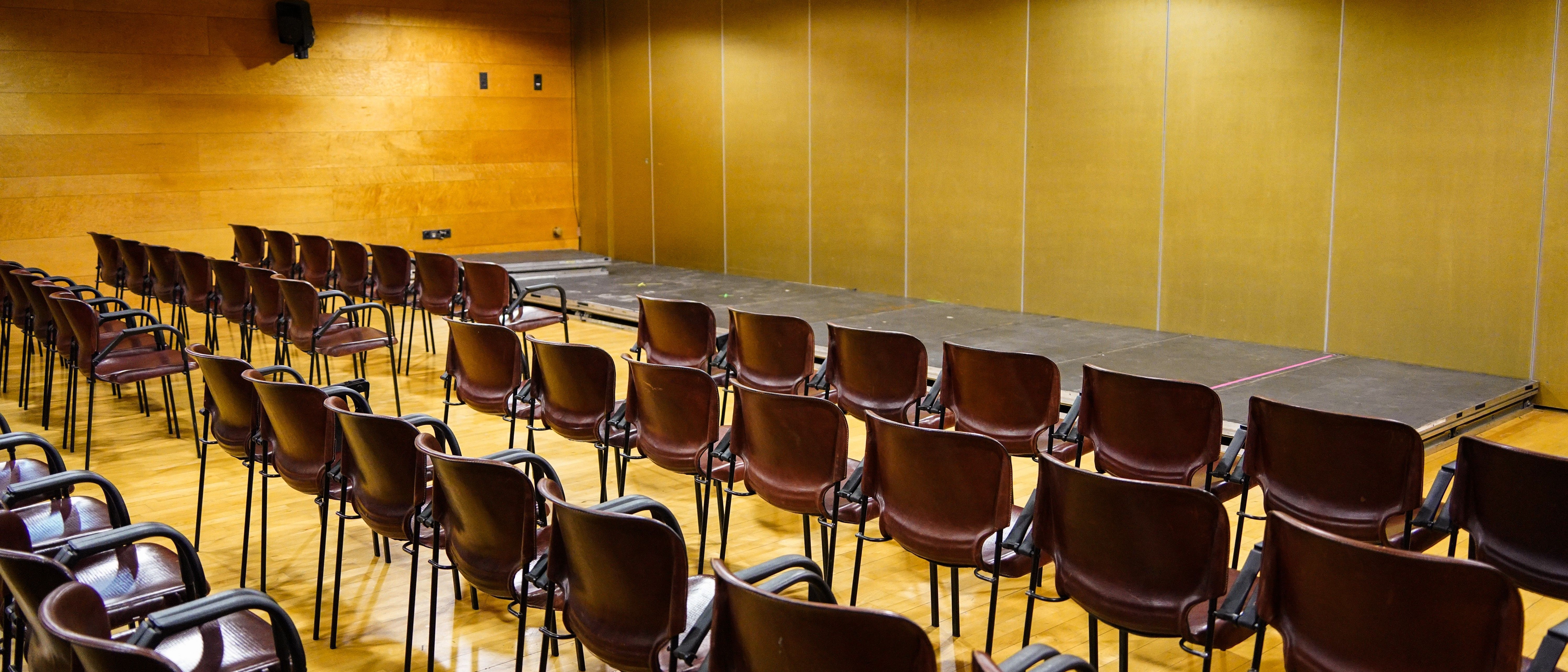 Rows of plain stacking chairs facing a small stage in a wood-panelled hall